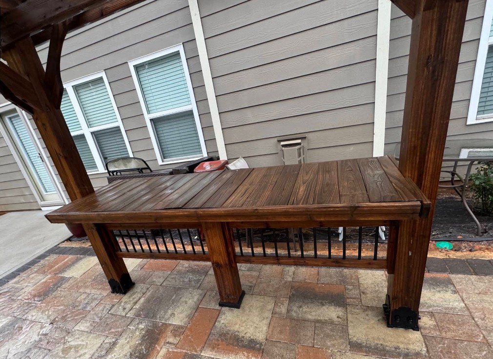 Dark-stained outdoor wood bar counter and posts on a stone patio beside a home