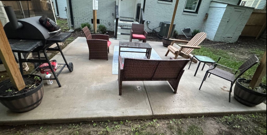 Residential backyard concrete patio with outdoor seating, grill, and brick home exterior.